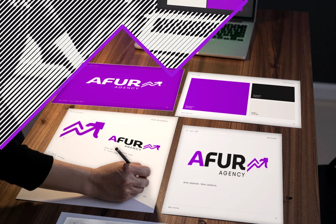 Dizayner masaya düzülmüş AFUR-⁠un brendbook vərəqləri üzərində karandaşla eskiz çəkir; noutbukda brendbook açıqdır.